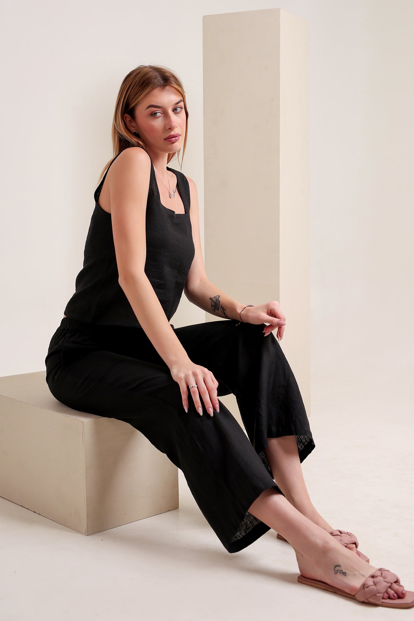 Woman wearing a black linen sleeveless top and pants set sitting on a white surface.