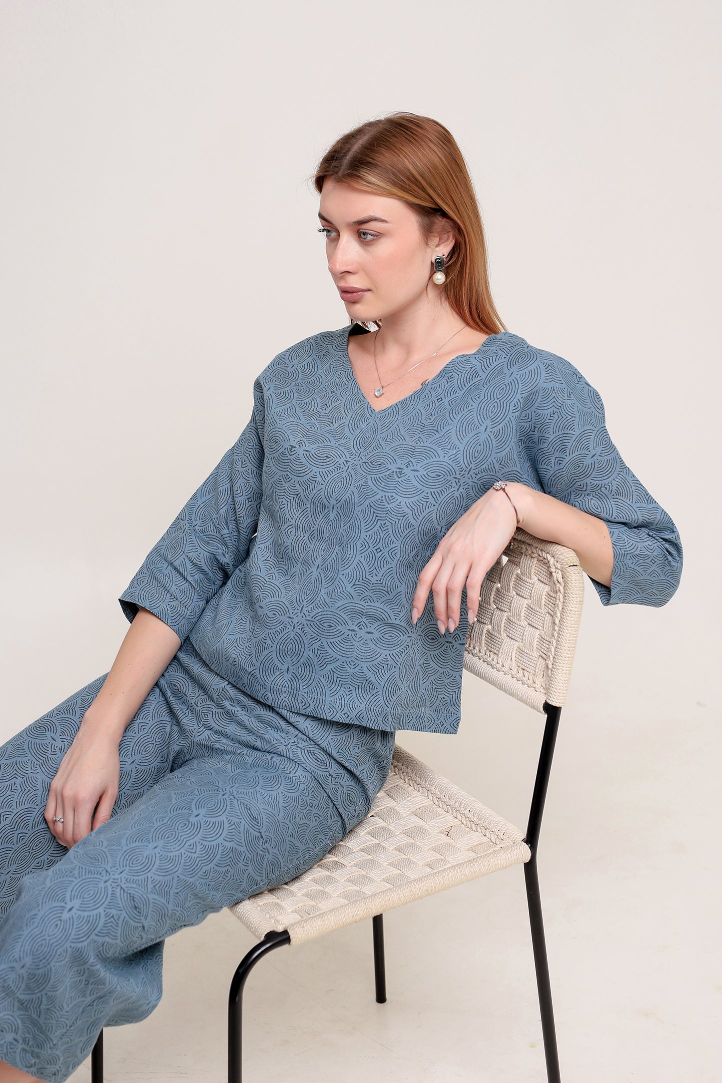 Woman wearing a blue textured linen top and pants set sitting on a chair against a white background.
