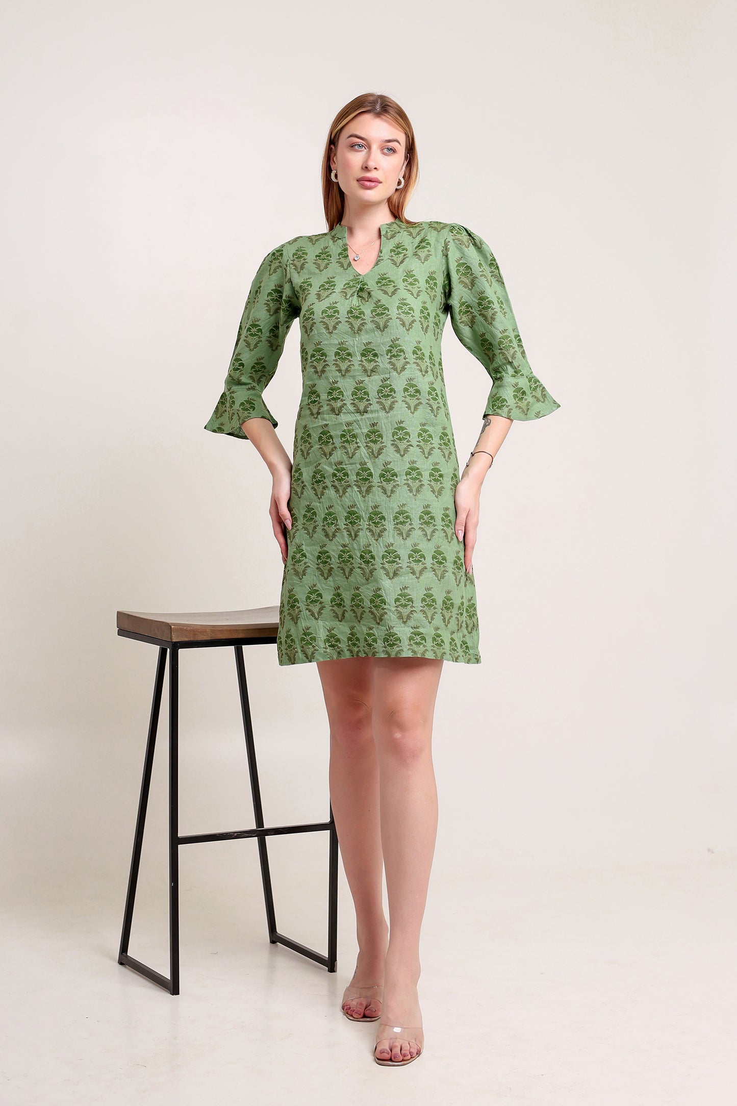 Woman wearing a green patterned linen mini dress standing next to a black stool on a white background.