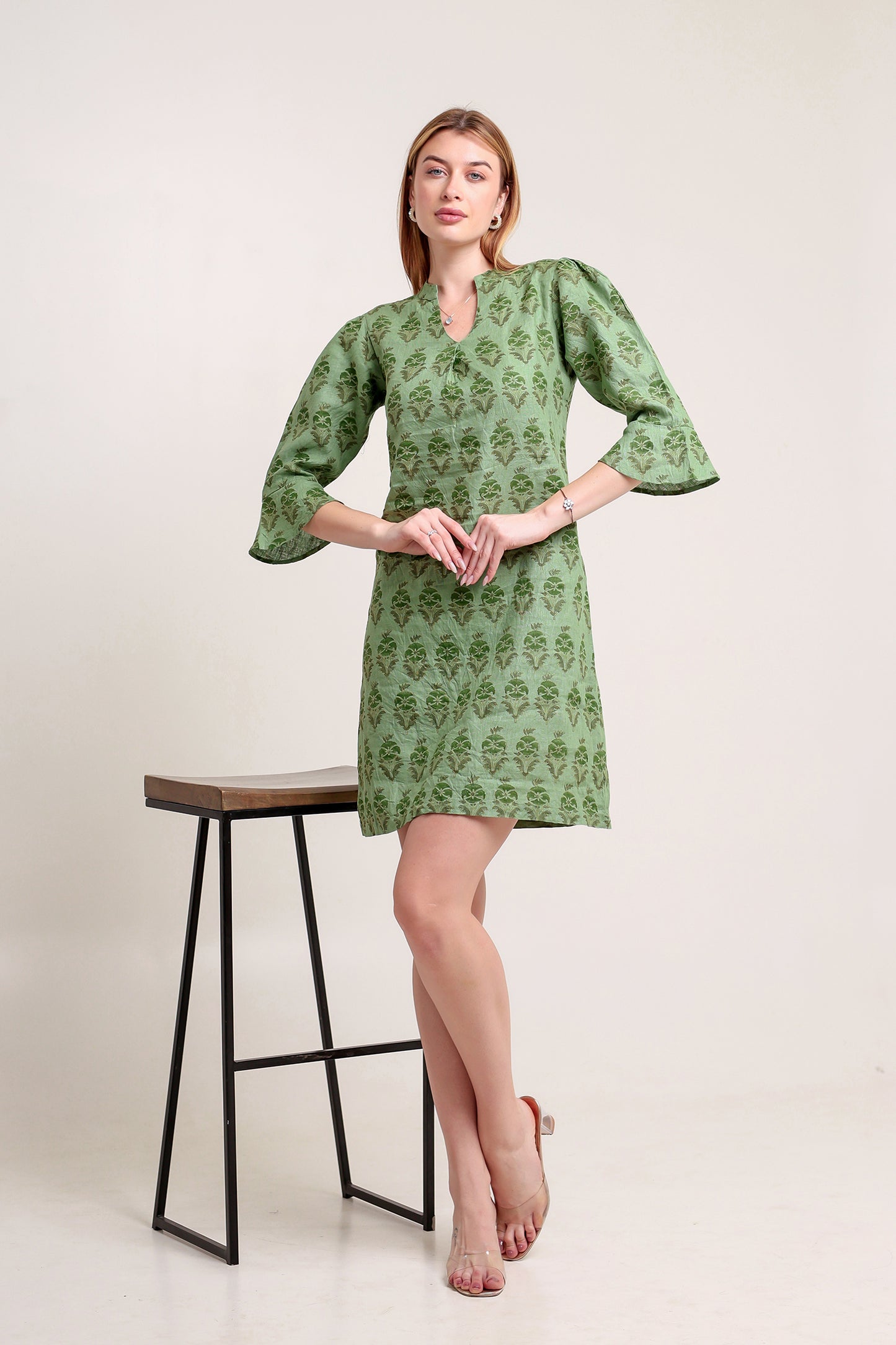Woman wearing a green patterned linen mini dress standing next to a black stool on a white background.