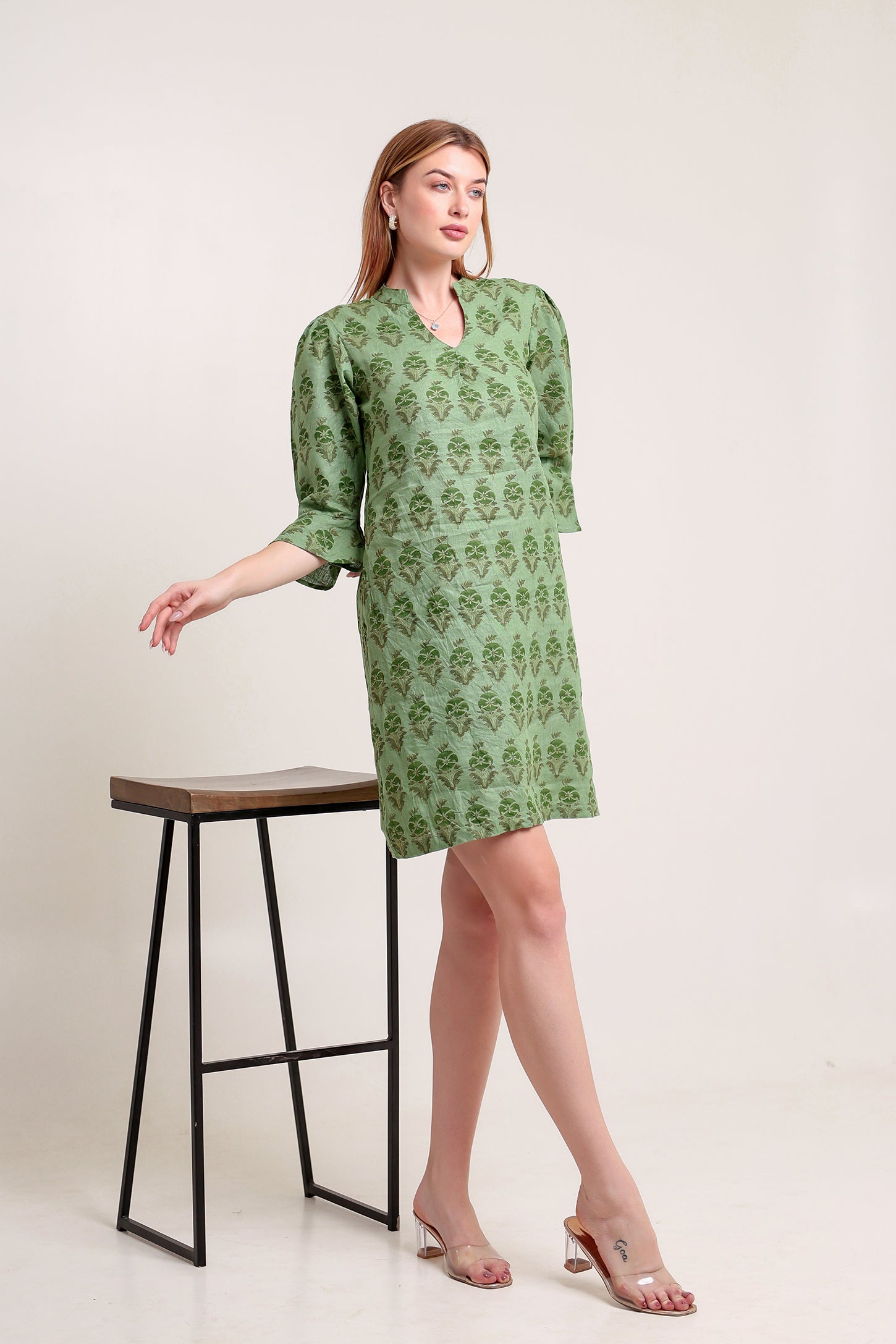 Woman wearing a green patterned linen mini dress standing next to a black stool on a white background.