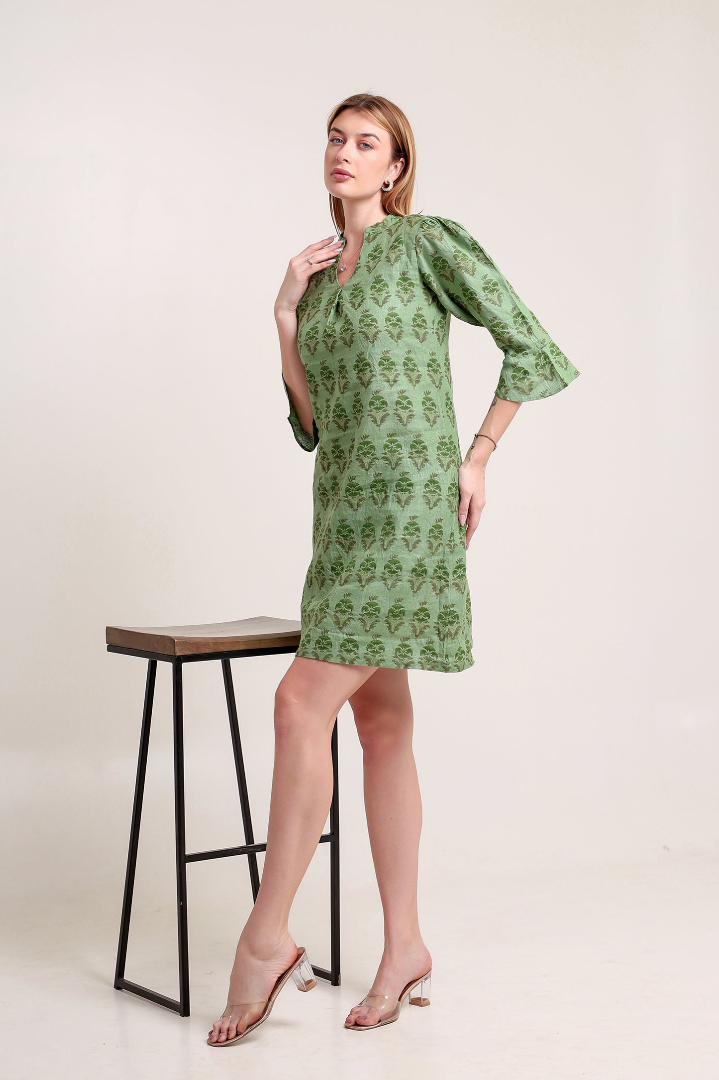 Woman wearing a green patterned linen mini dress standing next to a black stool on a white background.