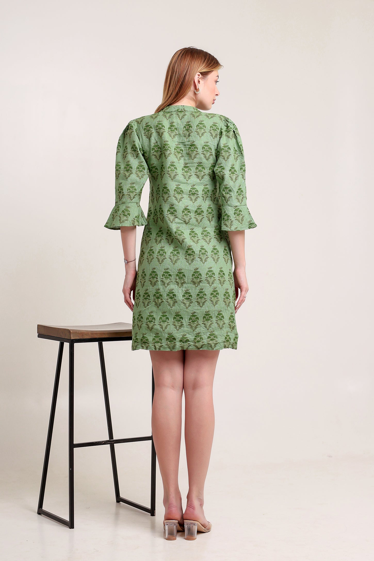 Woman wearing a green patterned linen mini dress standing next to a black stool on a white background.