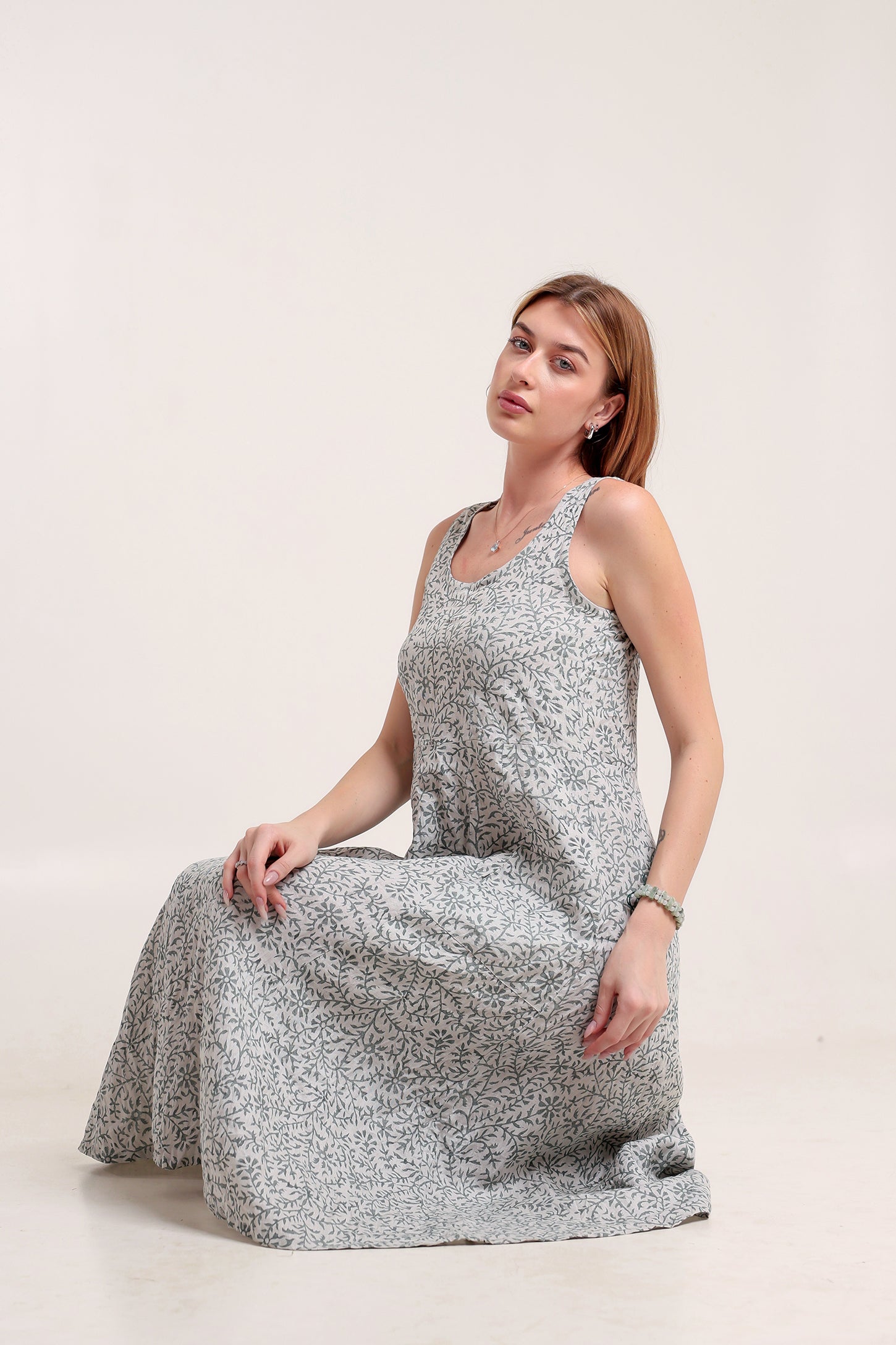 Woman wearing a sleeveless floral linen floral maxi dress sitting against a white background.
