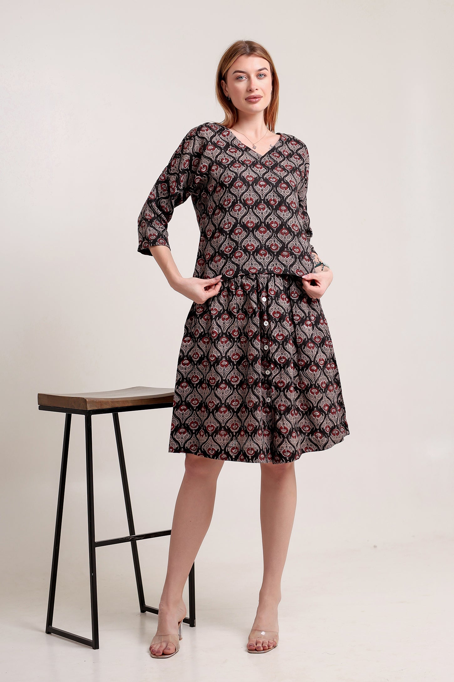 Woman wearing a stylish linen skirt and top set with patterns, standing next to a stool against a white background.
