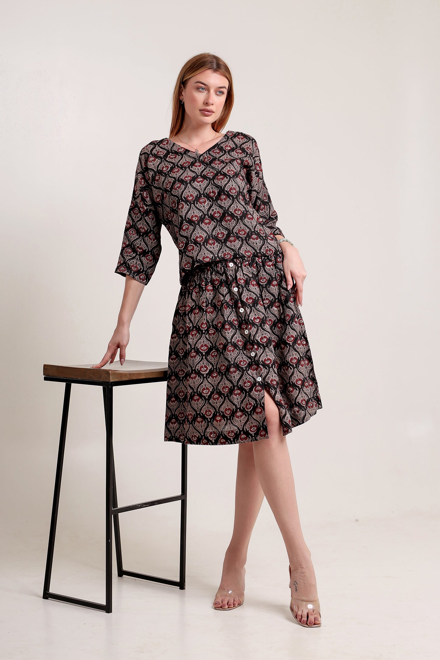 Woman wearing a patterned linen skirt and top set standing beside a stool against a plain background.
