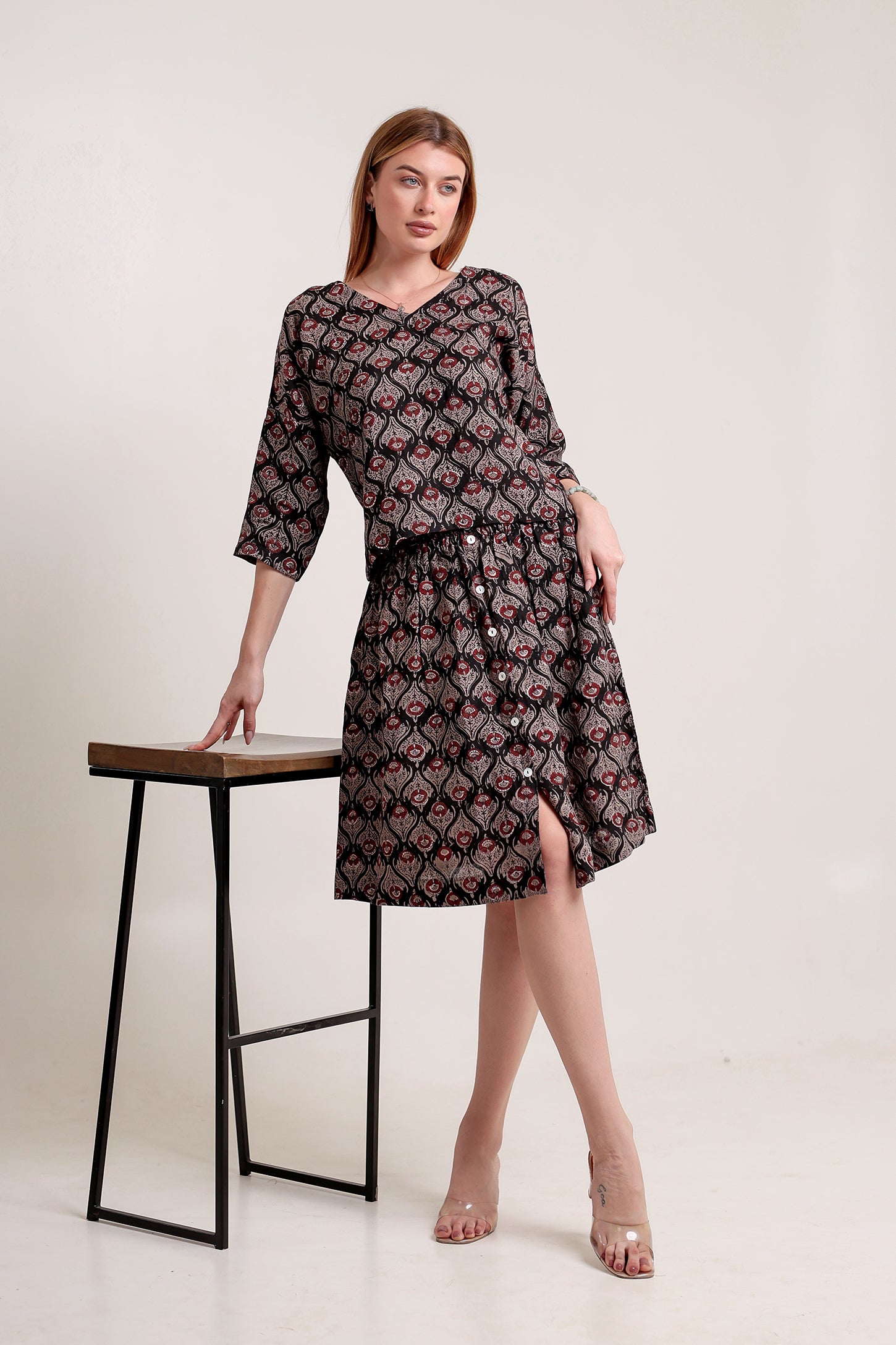 Woman wearing a patterned linen skirt and top set standing beside a stool against a plain background.
