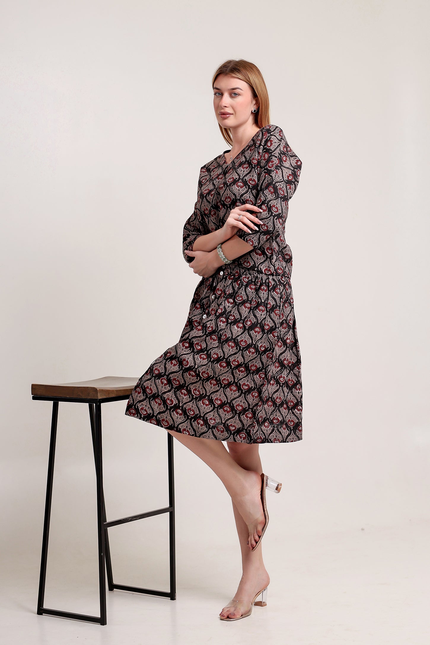 Woman wearing a patterned linen skirt and top set standing beside a stool against a white background.