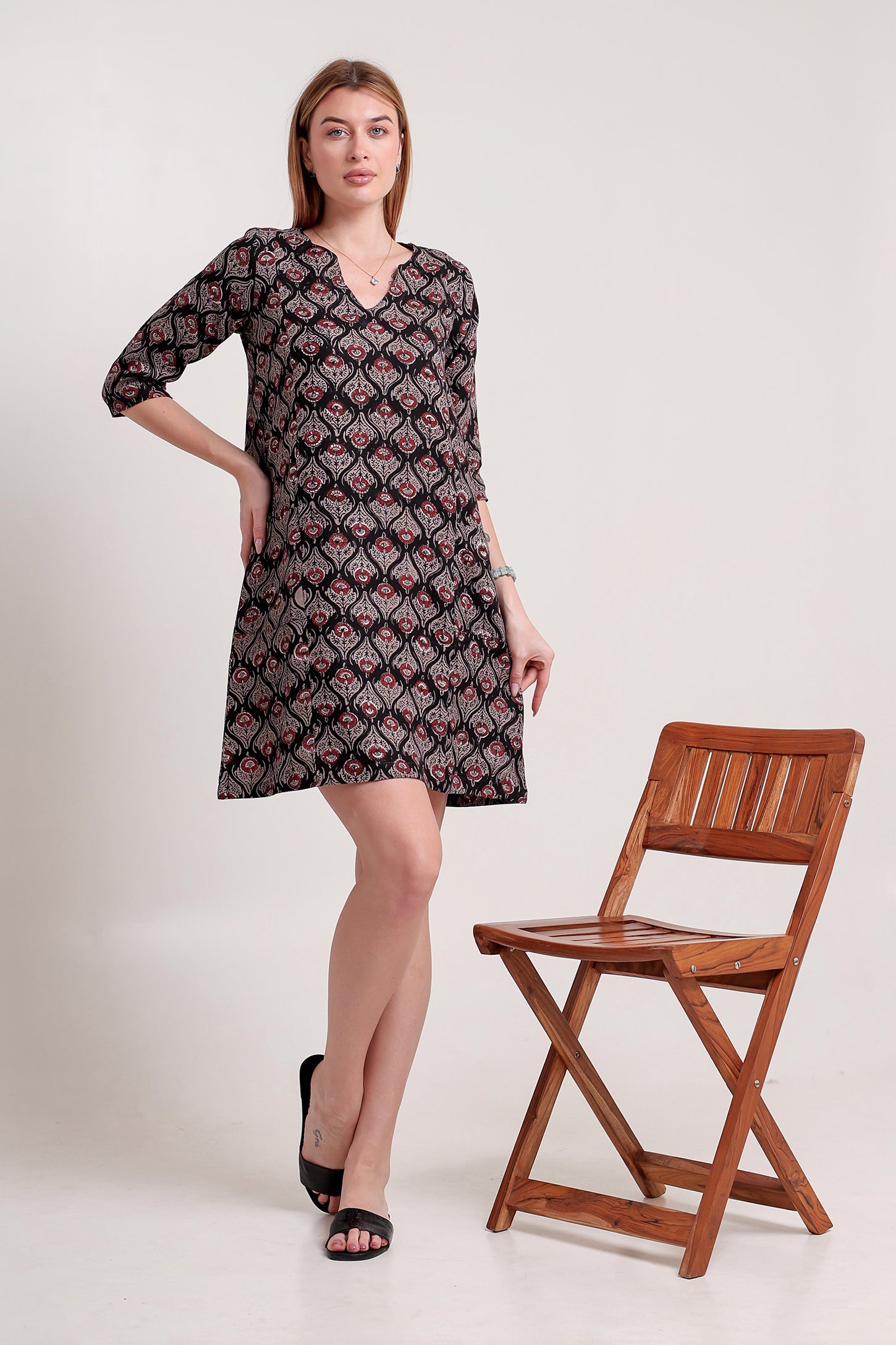 Woman wearing a patterned linen mini dress standing next to a wooden chair on a white background.