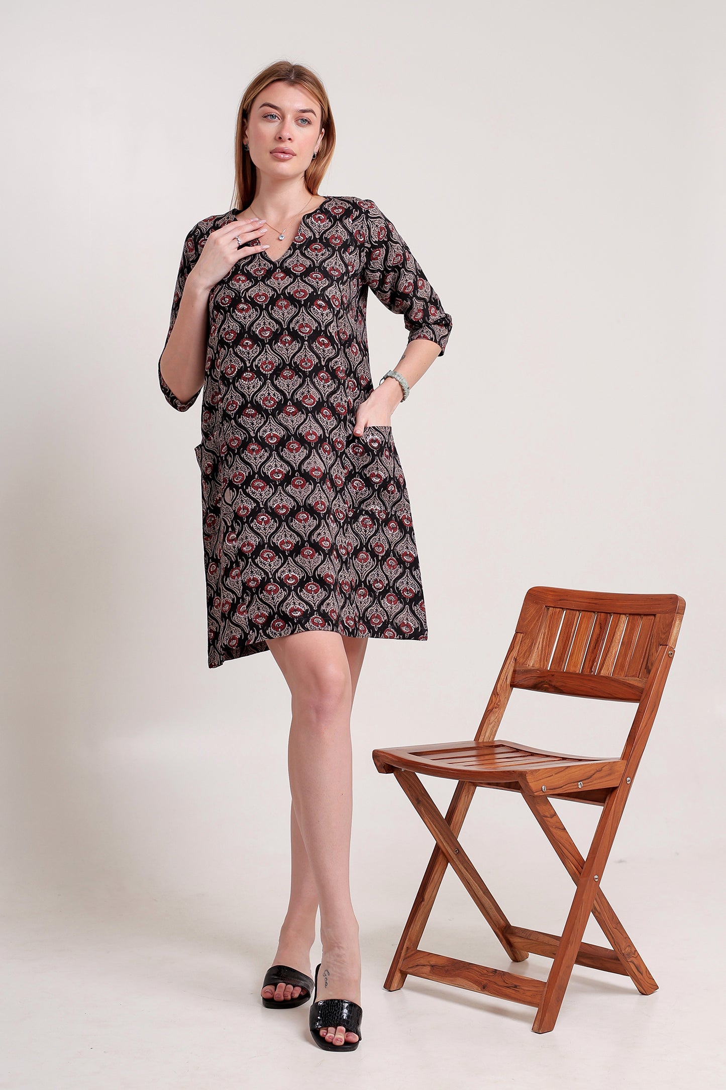 Woman wearing a patterned linen mini dress standing next to a wooden chair on a white background.
