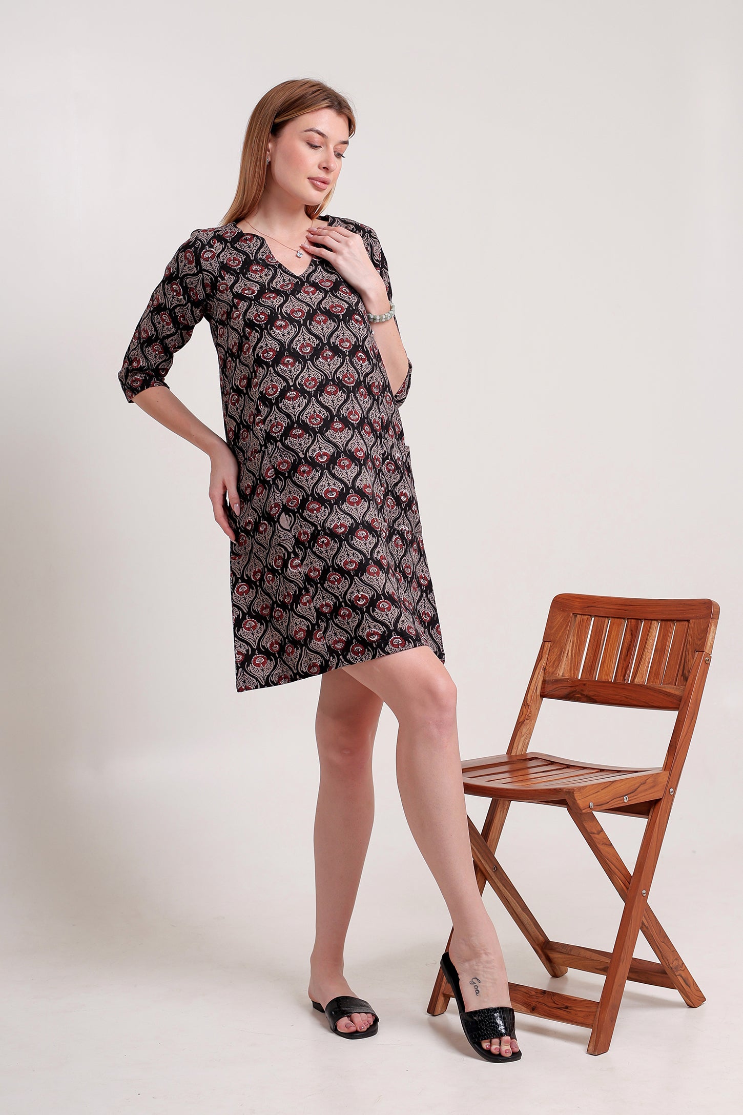 Woman wearing a patterned linen mini dress standing next to a wooden chair on a white background.