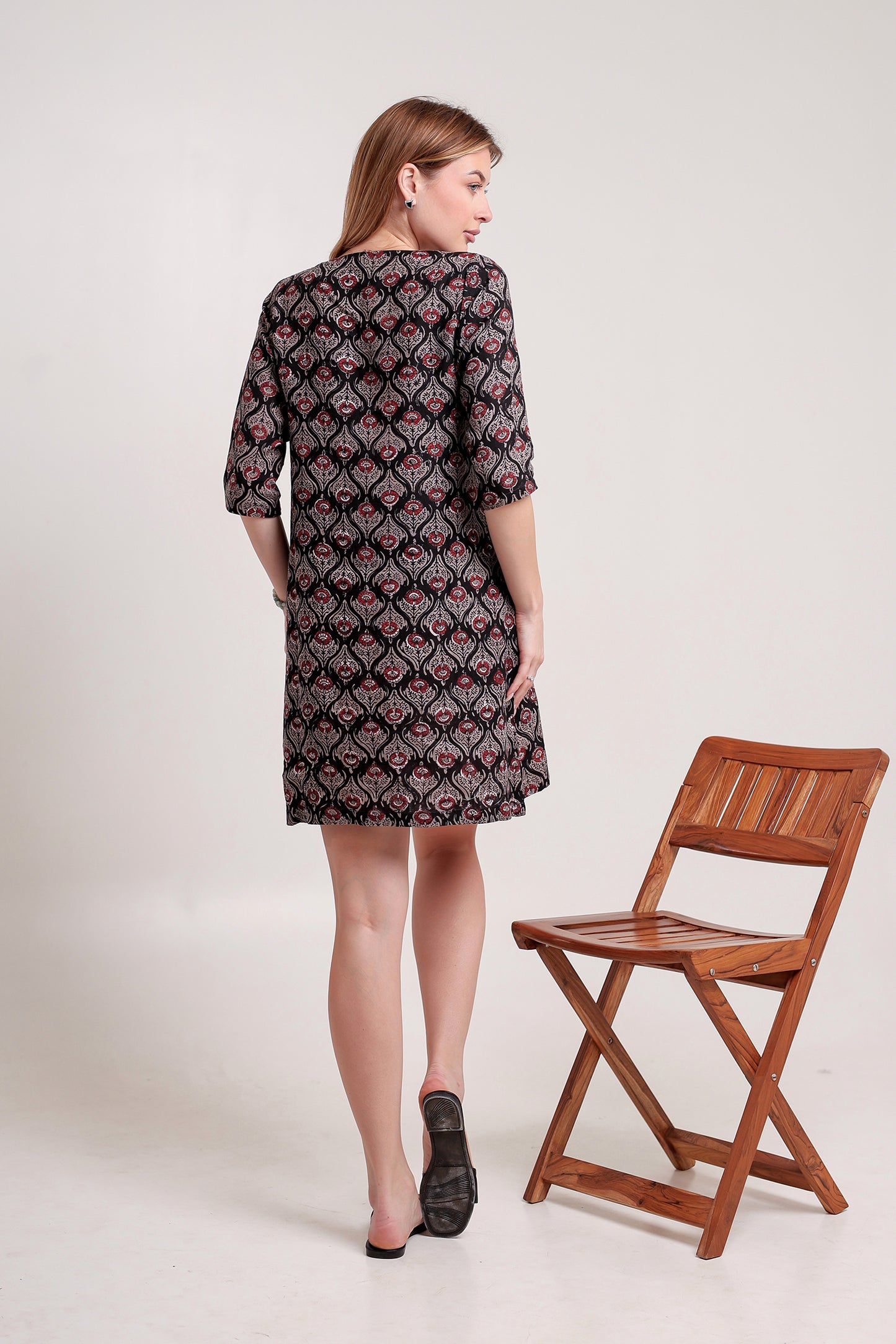 Woman wearing a patterned linen mini dress standing next to a wooden chair on a white background.