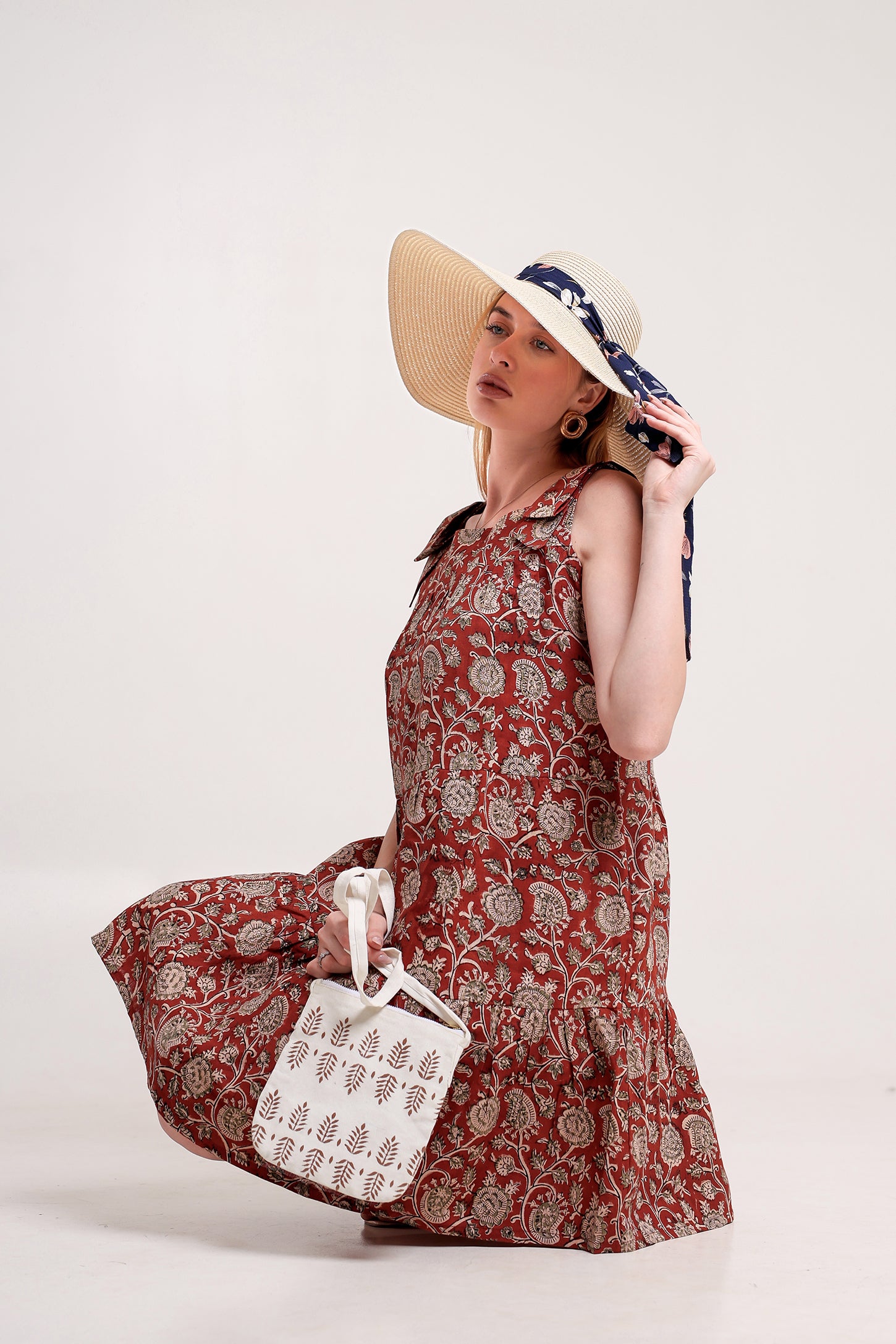 Woman wearing a red floral linen floral mini dress with a white bag and straw hat against a white background.