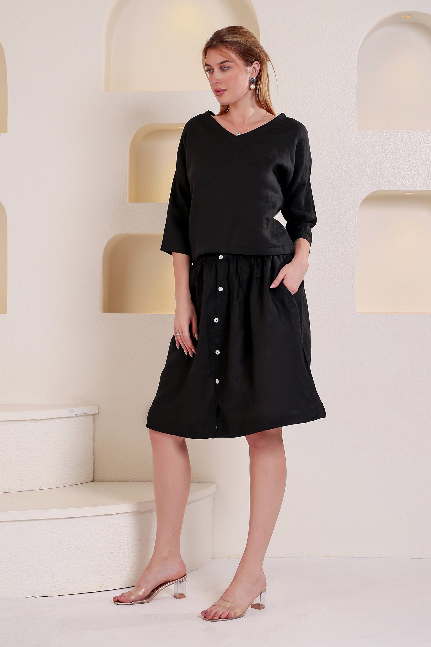 Woman wearing a black buttoned linen top and skirt set standing in a minimalistic setting.