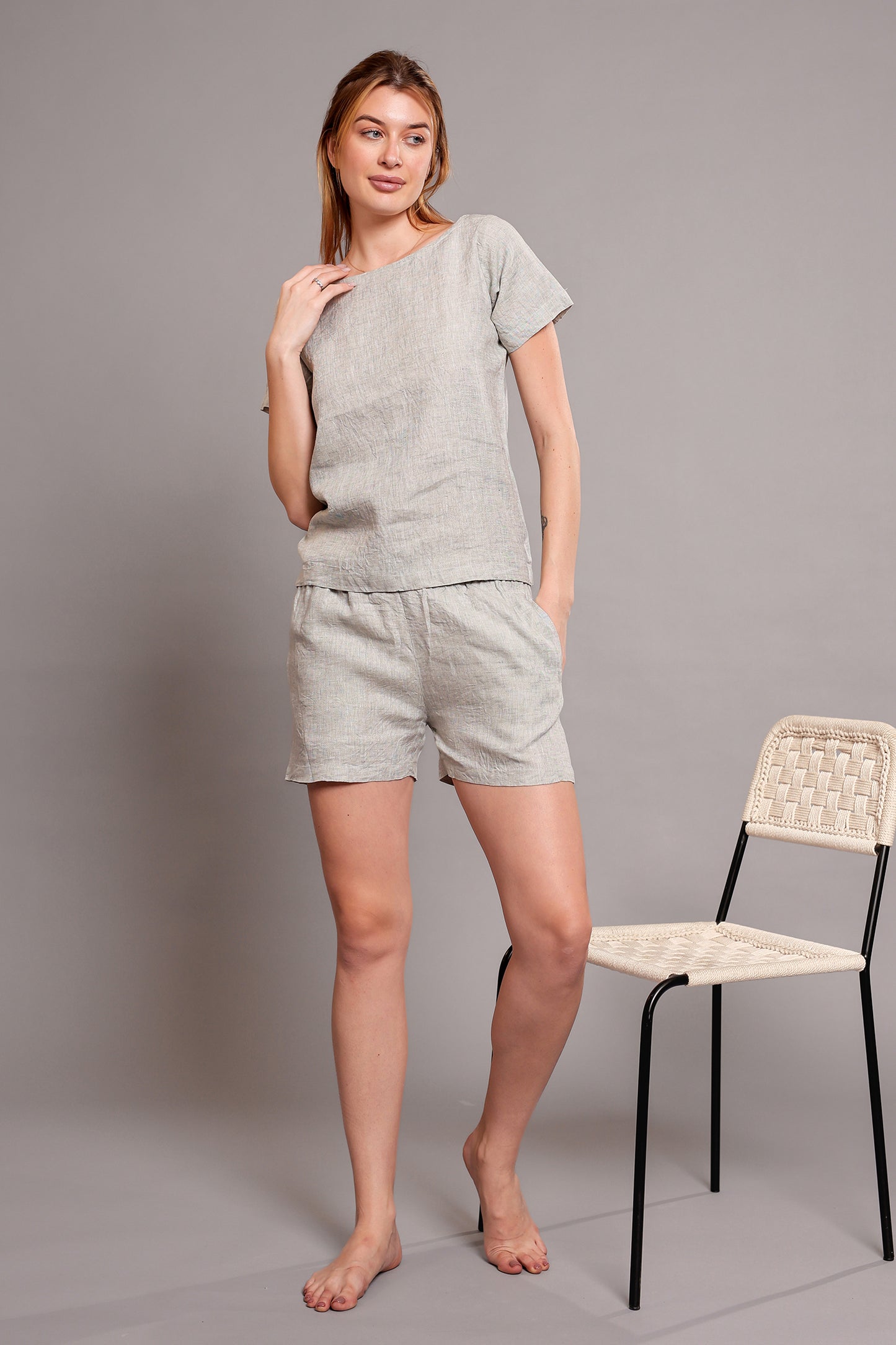 Woman wearing a light gray linen top and shorts set standing next to a chair on a plain background.