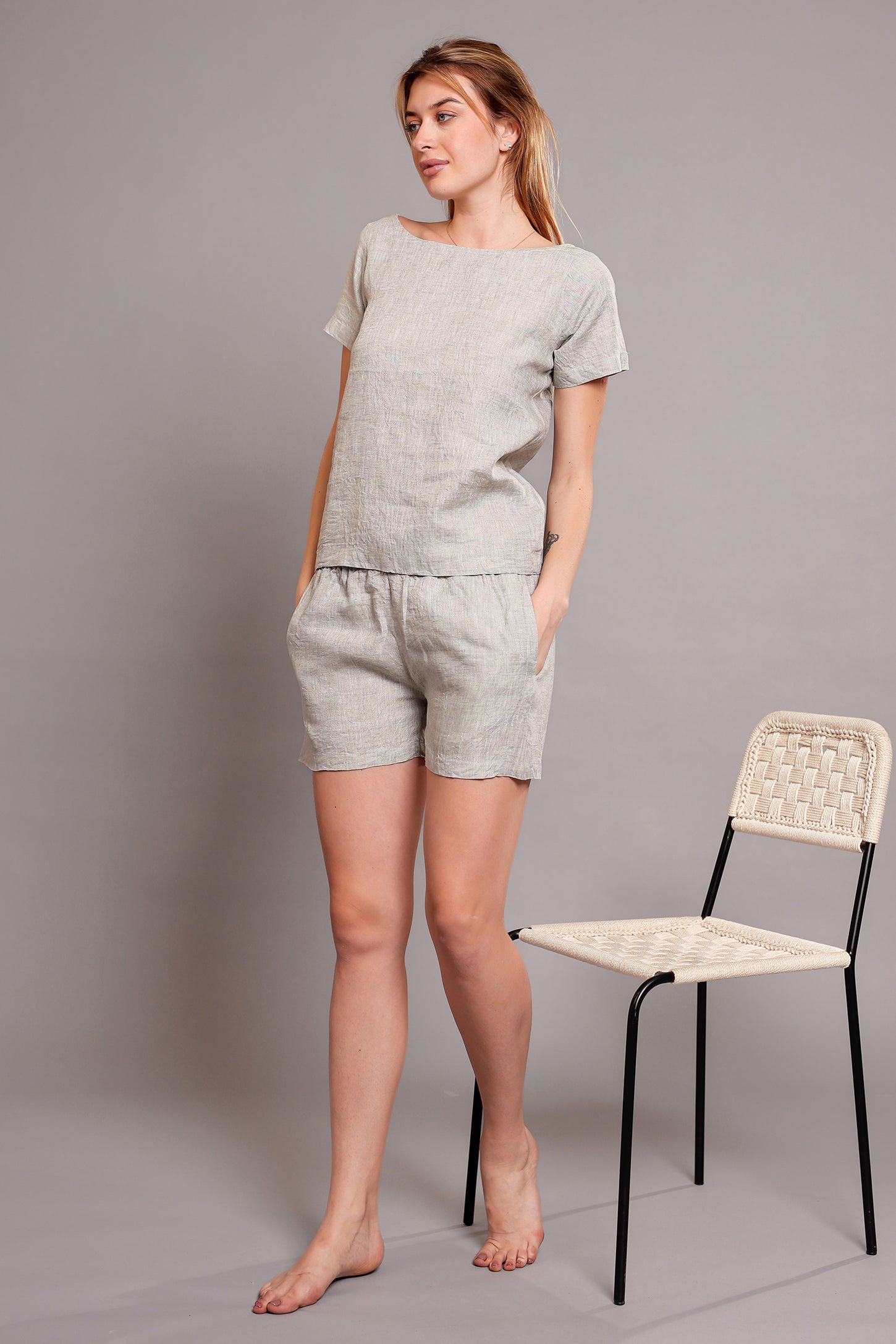 Woman wearing a light gray linen top and shorts set standing next to a chair against a plain background.