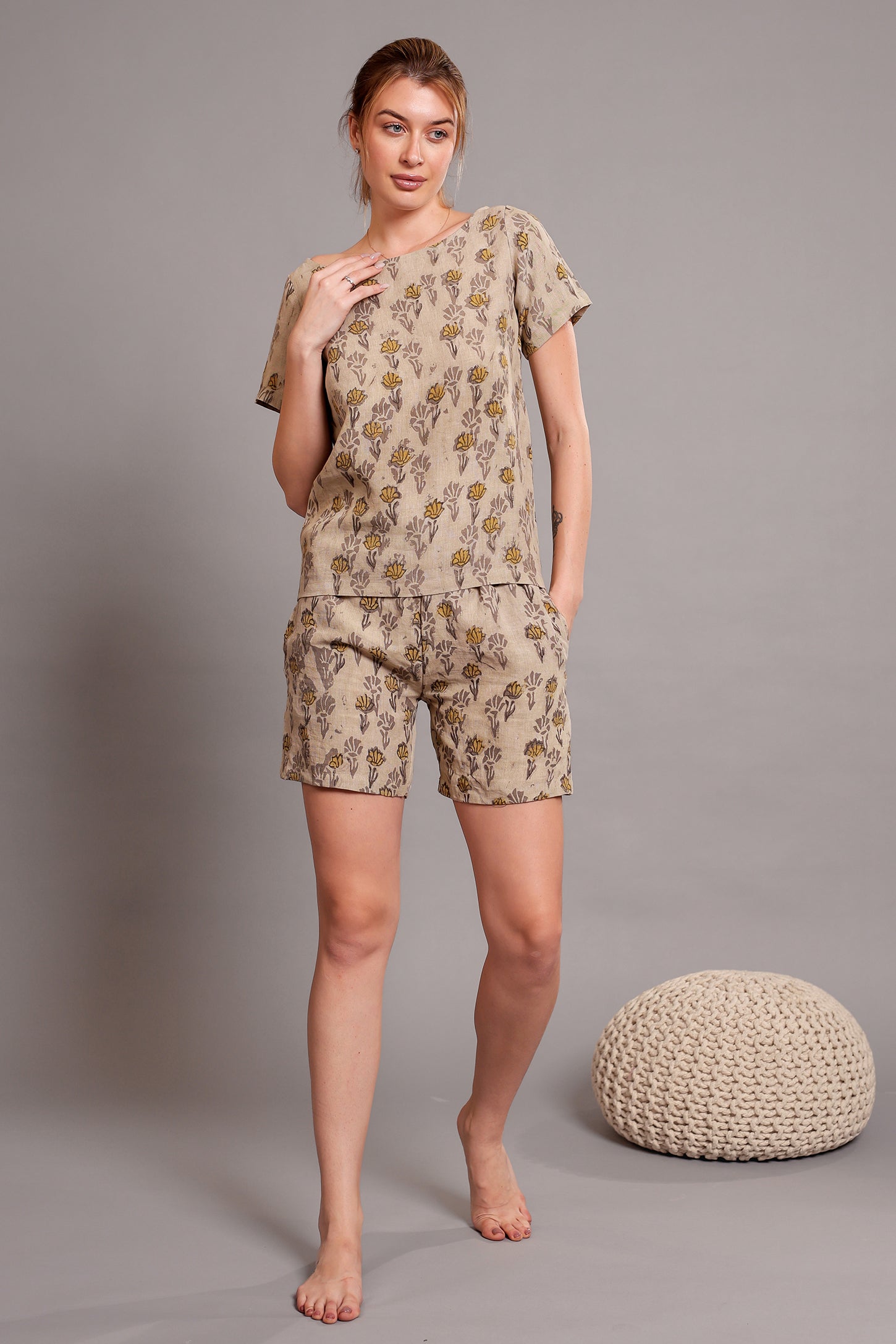 Woman wearing a beige linen floral top and shorts pajama set standing against a gray background with a textured stool.