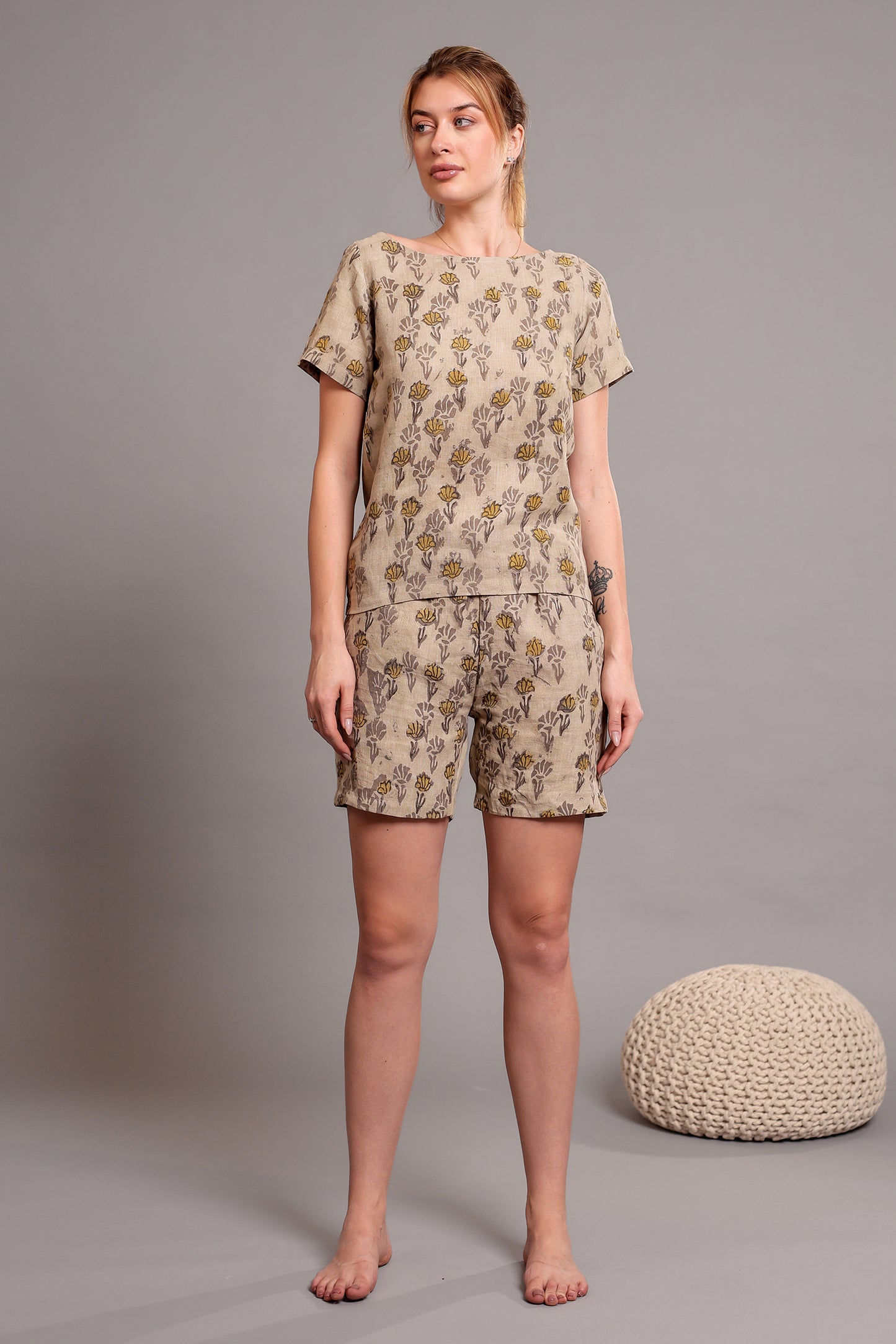 Woman wearing a beige patterned linen floral top and shorts set standing against a gray background with a textured round object.