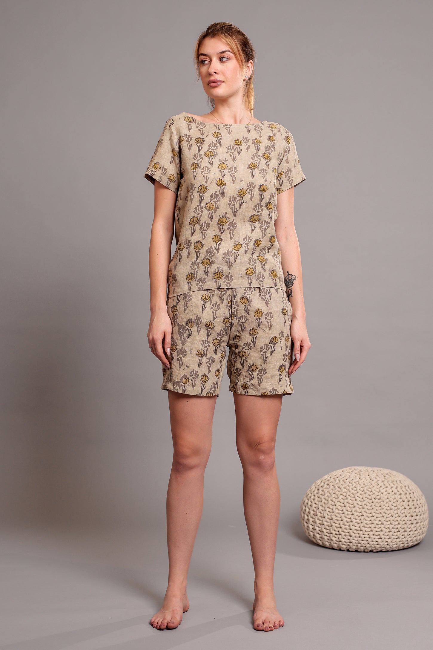 Woman wearing a beige patterned linen floral top and shorts set standing against a gray background with a textured round object.