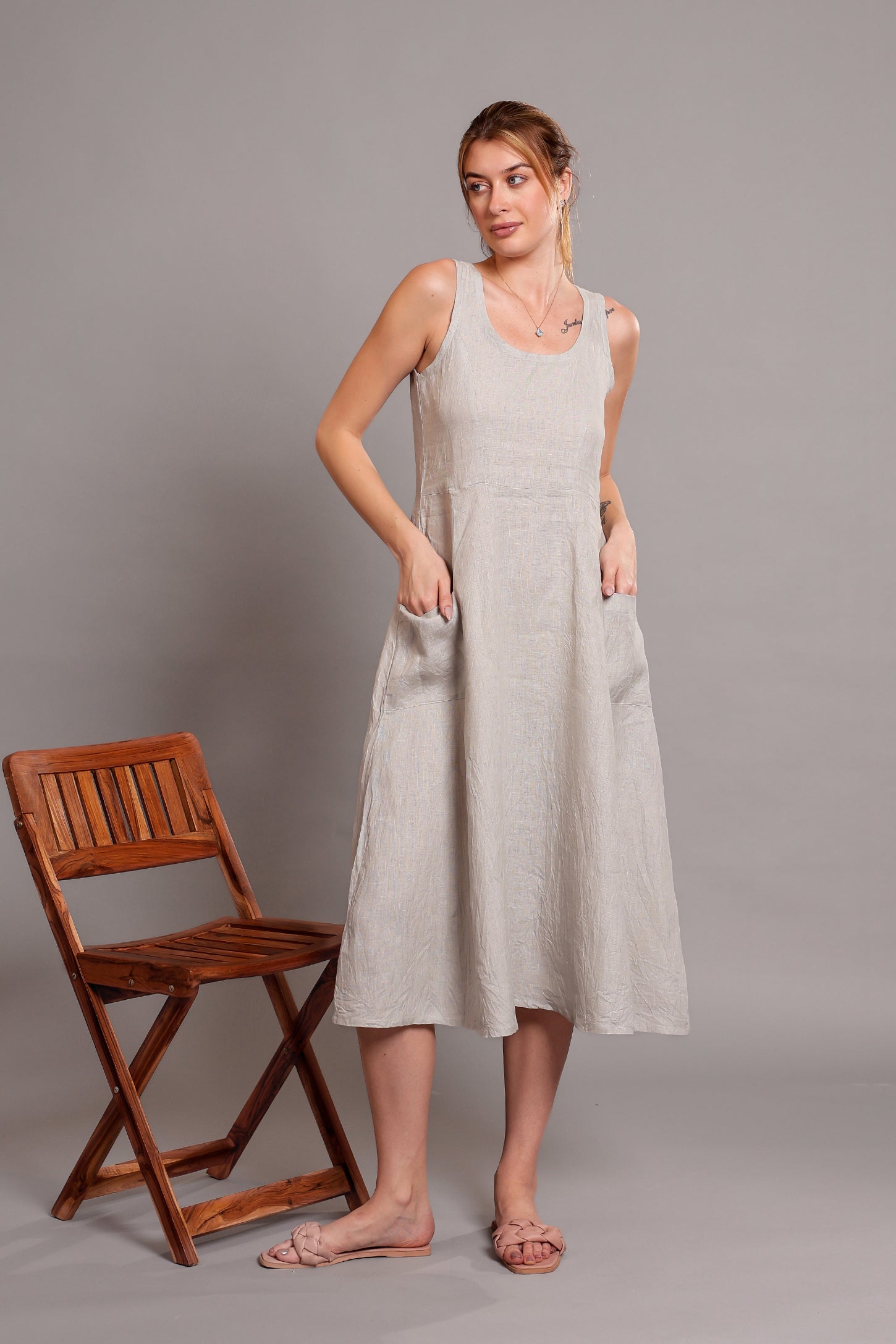 Woman wearing a light-colored linen sleeveless dress standing next to a wooden chair on a gray background.