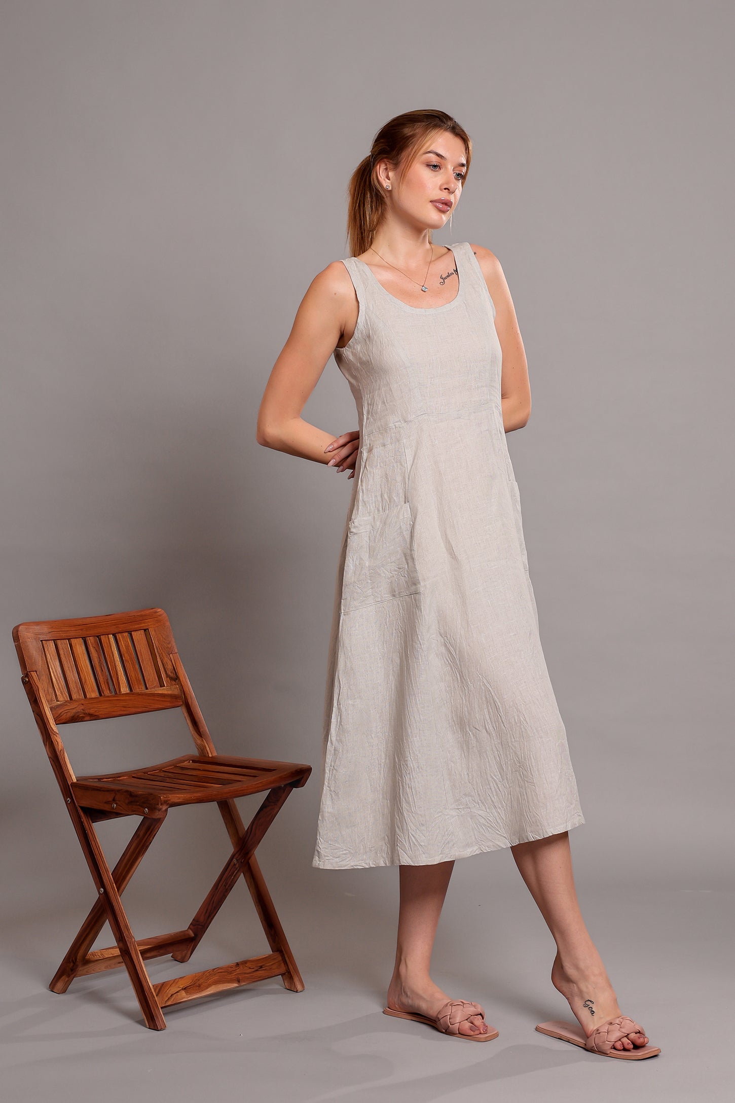 Woman wearing a light-colored linen sleeveless dress standing beside a wooden chair against a gray background.