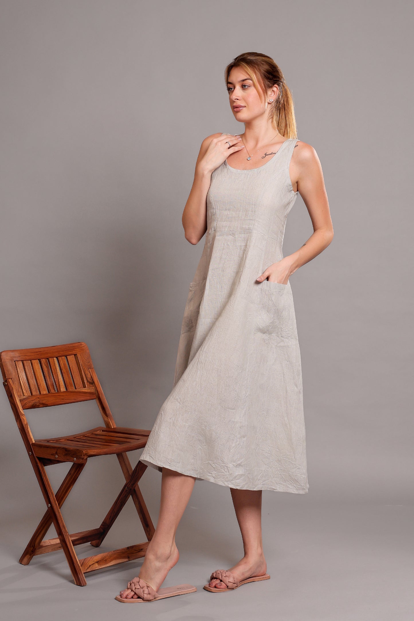 Woman wearing a light gray linen sleeveless dress standing beside a wooden chair on a plain background.