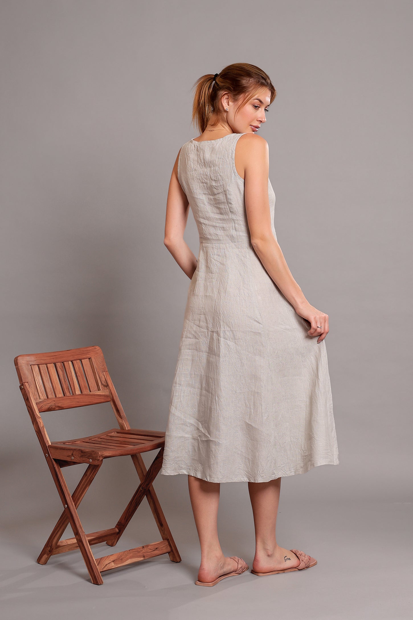 Woman wearing a light-colored linen midi dress standing beside a wooden chair against a gray background.