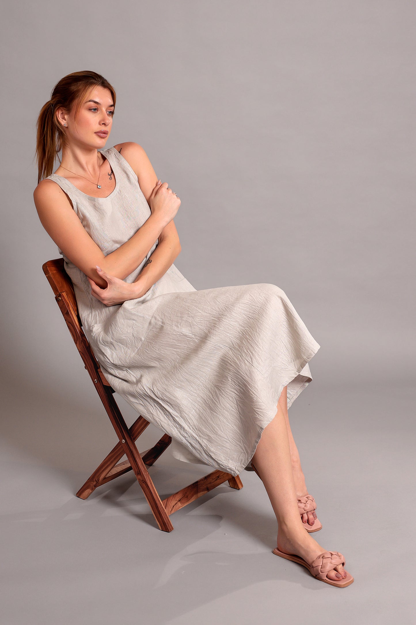 Woman wearing a beige linen midi dress seated on a wooden chair against a gray background.