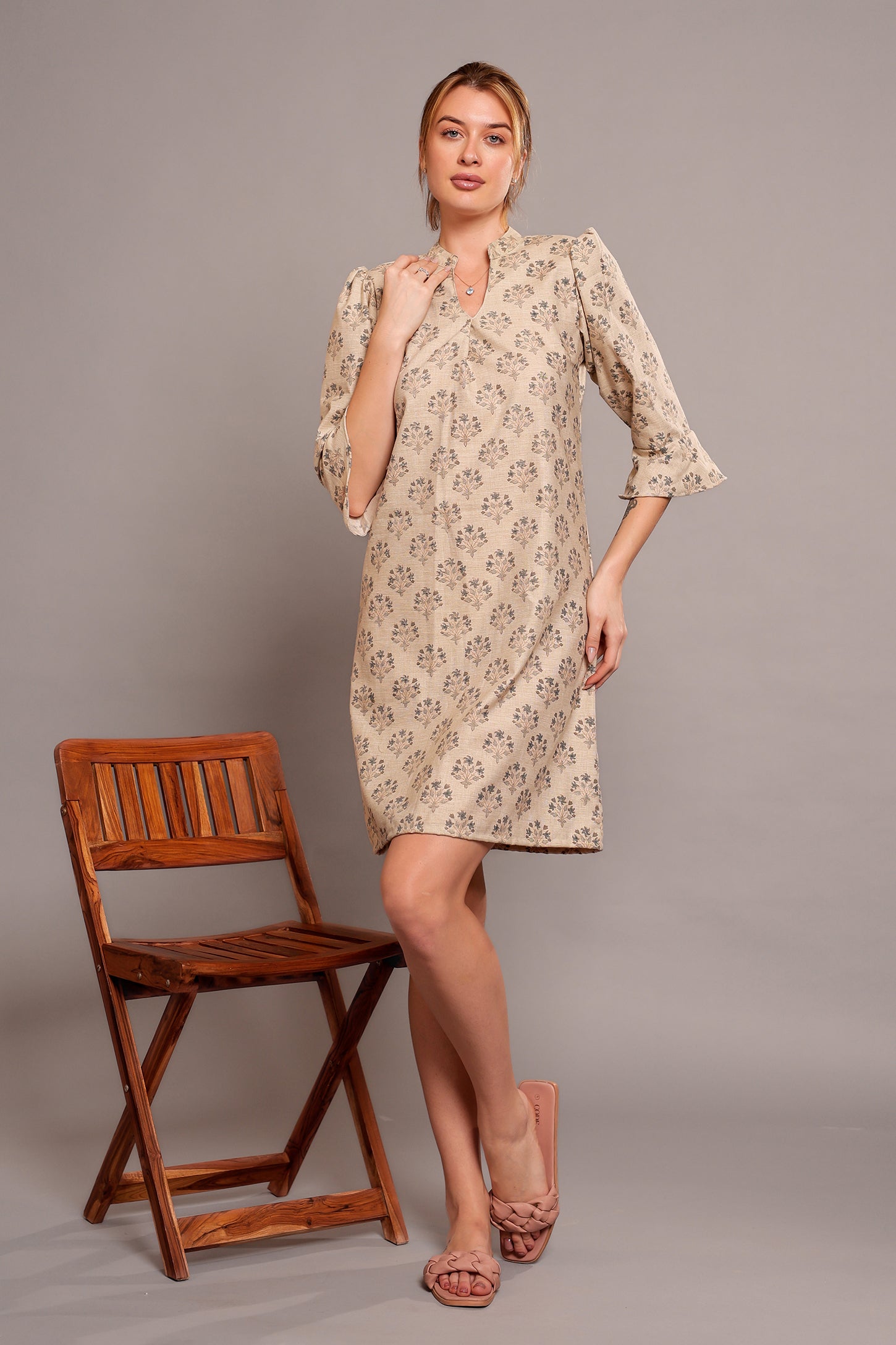 Woman wearing a linen floral dress standing beside a wooden chair against a gray background.