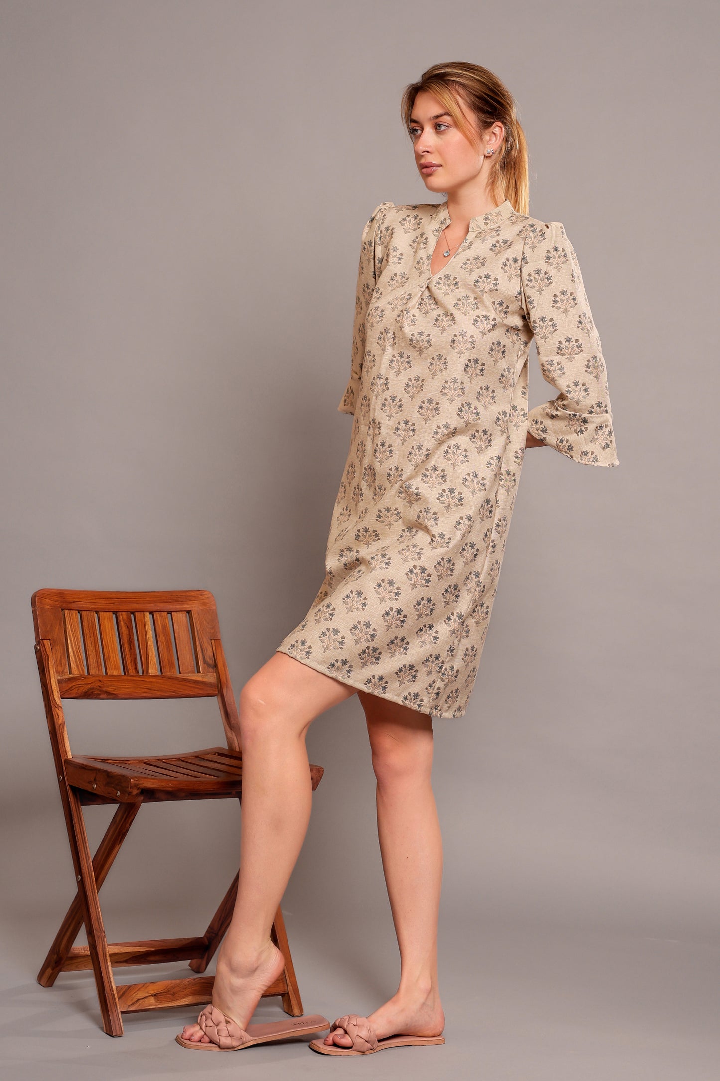 Woman wearing a linen floral dress standing beside a wooden chair against a gray background.