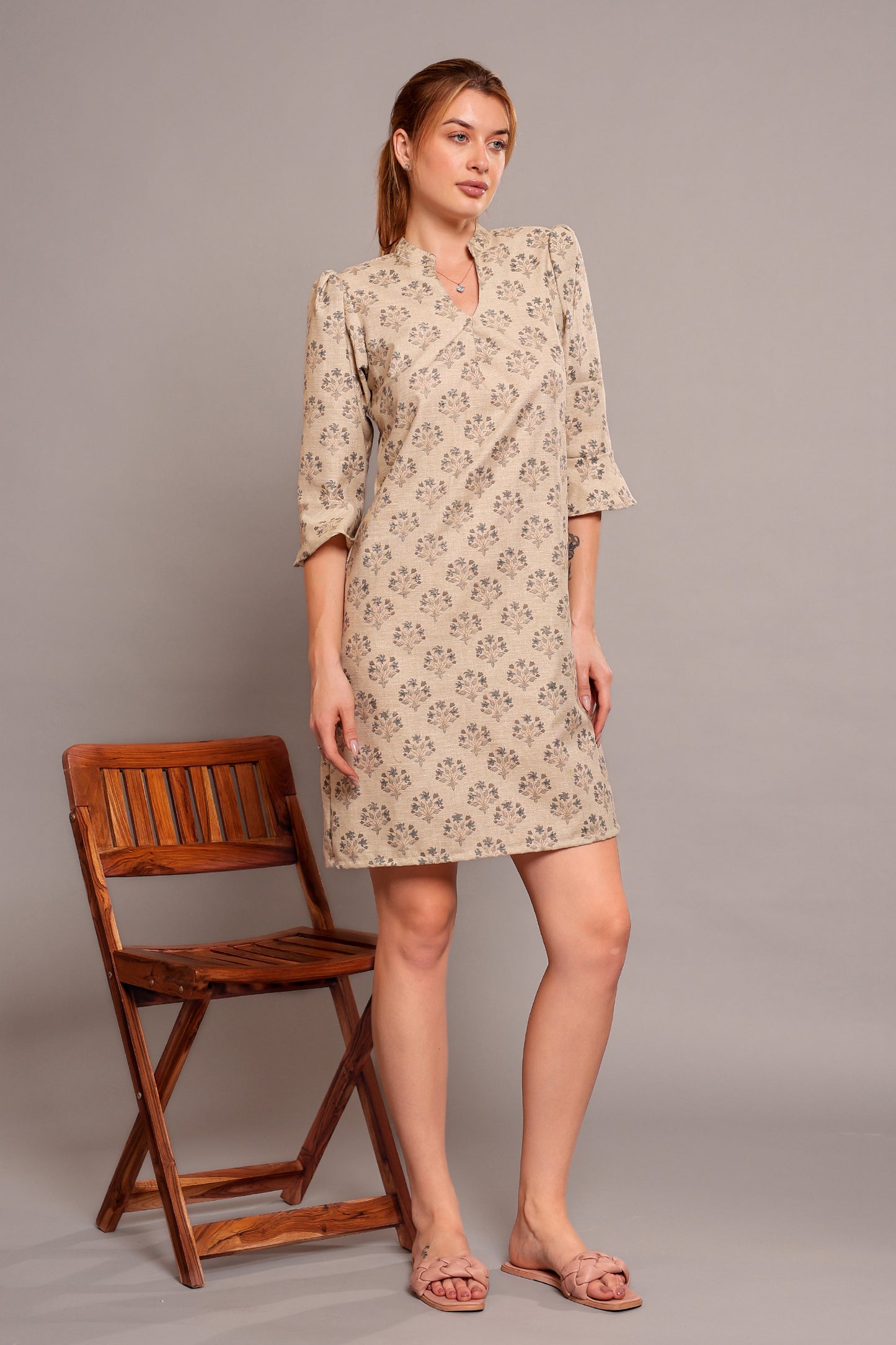 Woman wearing a patterned linen floral dress standing beside a wooden chair on a gray background.