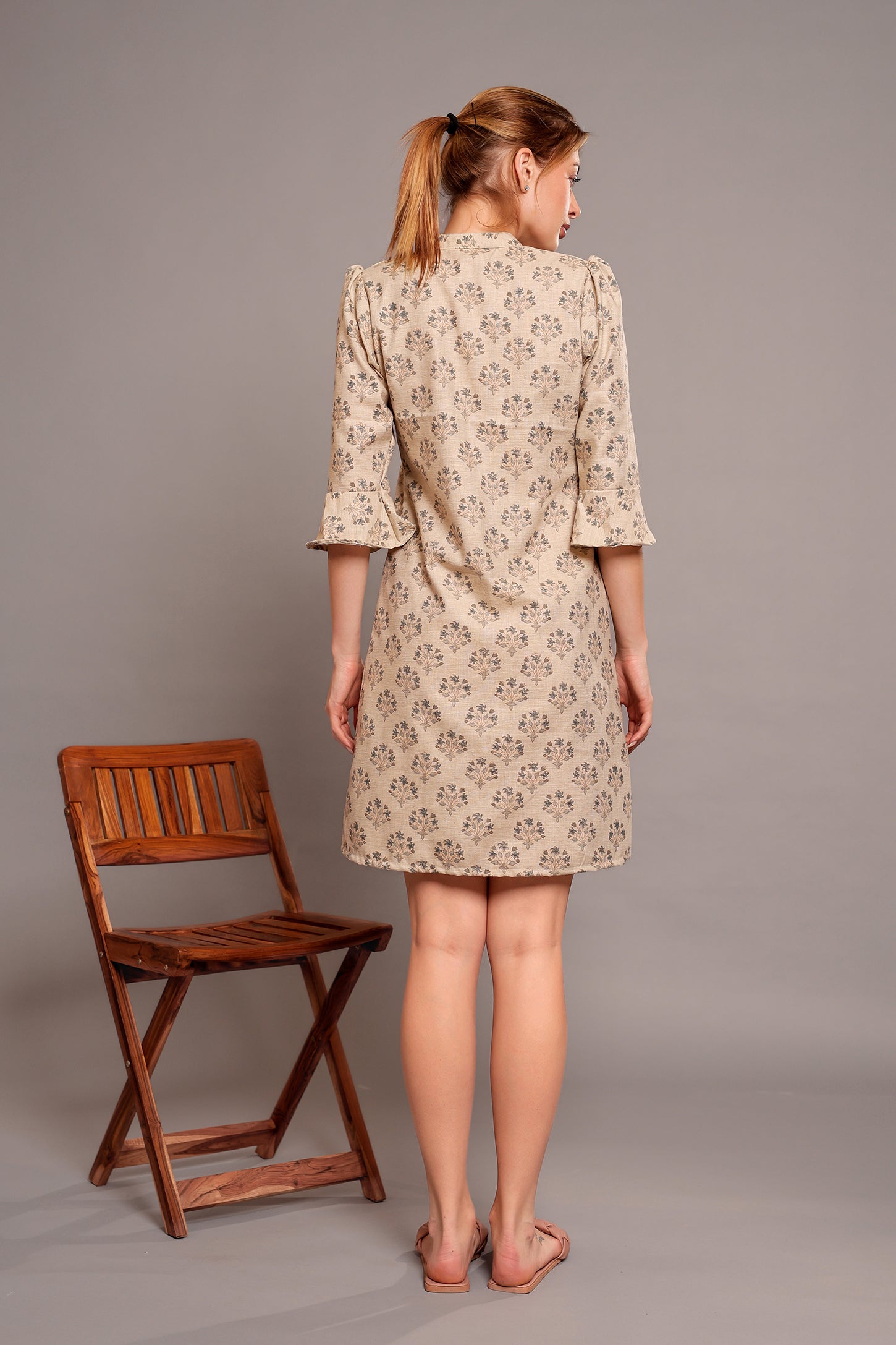 Woman wearing a patterned linen floral dress standing next to a wooden chair on a plain background.